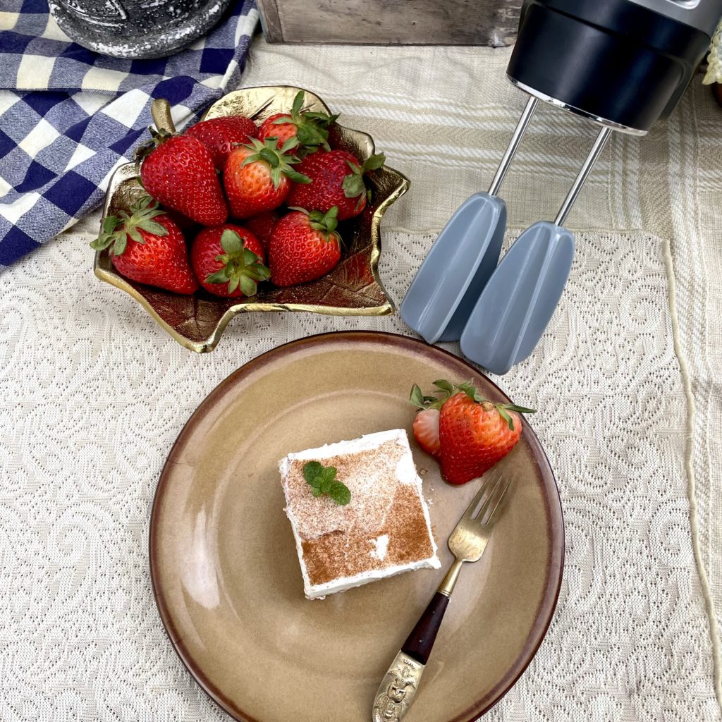 A sponge cake soaked with three milks and then topped with whipped cream. The plate has a serving with a strawberry sliced up. In the background, there is a bowl of strawberries, the Hamilton Beach Mixer and some planters for aesthetics.